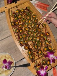 Wooden platter of restaurant-style sushi bites: rows of crispy rice topped with thinly sliced fish, jalapeño rings, spicy orange mayo and drizzled dark sauce, garnished with purple orchids and pickled ginger while chopsticks lift a piece.