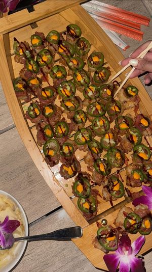 Wooden platter of restaurant-style sushi bites: rows of crispy rice topped with thinly sliced fish, jalapeño rings, spicy orange mayo and drizzled dark sauce, garnished with purple orchids and pickled ginger while chopsticks lift a piece.