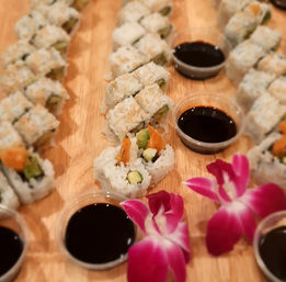 Close-up sushi platter on a wooden board: rice-wrapped rolls with avocado and orange filling, small soy sauce cups, and bright pink orchid garnishes