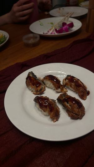 Five pieces of grilled eel (unagi) nigiri sushi drizzled with glossy sauce on a white plate, set on a wooden dining table with chopsticks, edamame and plates in the background.
