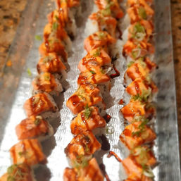 Mouthwatering close-up of three rows of spicy salmon sushi rolls on a metal platter, topped with creamy spicy mayo, sweet eel sauce drizzle and chopped scallions.