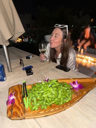 Playful woman with white sunglasses on her head puckering while holding a wine glass at a nighttime rooftop patio table, wooden boat of edamame with orchid garnish in the foreground and a glowing fire feature in the background.