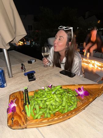 Playful woman with white sunglasses on her head puckering while holding a wine glass at a nighttime rooftop patio table, wooden boat of edamame with orchid garnish in the foreground and a glowing fire feature in the background.