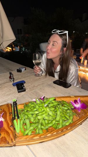 Playful woman with white sunglasses on her head puckering while holding a wine glass at a nighttime rooftop patio table, wooden boat of edamame with orchid garnish in the foreground and a glowing fire feature in the background.