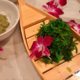 Fresh green seaweed salad garnished with a magenta orchid on a small wooden sushi-boat platter, served next to a bowl of wasabi