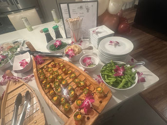 Indoor sushi catering table with wooden boat platter of jalapeño-topped sushi bites, bowl of edamame, seaweed salad, soy sauce bottles, chopsticks and stacked plates, decorated with purple orchid garnishes