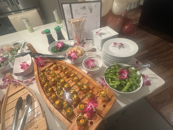 Indoor sushi catering table with wooden boat platter of jalapeño-topped sushi bites, bowl of edamame, seaweed salad, soy sauce bottles, chopsticks and stacked plates, decorated with purple orchid garnishes