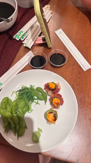 Japanese appetizer plate on a wooden table with edamame pods, seaweed salad, three tuna tartare sushi bites topped with small orange yolks, a dollop of wasabi, two soy sauce cups, chopstick wrappers and a sauce bowl.