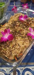 Glass baking dish of stir-fried noodles with shredded seaweed, carrots and cabbage, topped with bright purple orchid blossoms on a blue tiled table