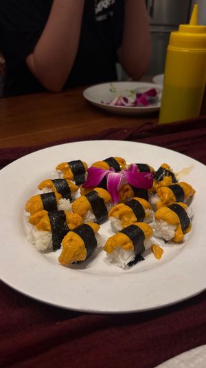 White plate of uni (sea urchin) nigiri sushi — rice topped with bright orange uni and nori, purple orchid garnish on a wooden dining table with a yellow squeeze bottle in the background.