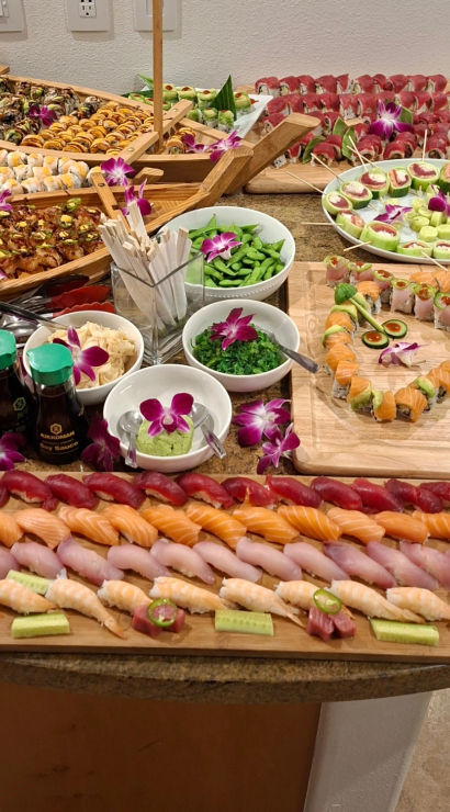 Vibrant sushi buffet and catering spread on wooden boards and boat platters with rows of nigiri, assorted maki rolls, cucumber bites, edamame, seaweed salad, wasabi and soy sauce bottles, orchid garnishes for a colorful party