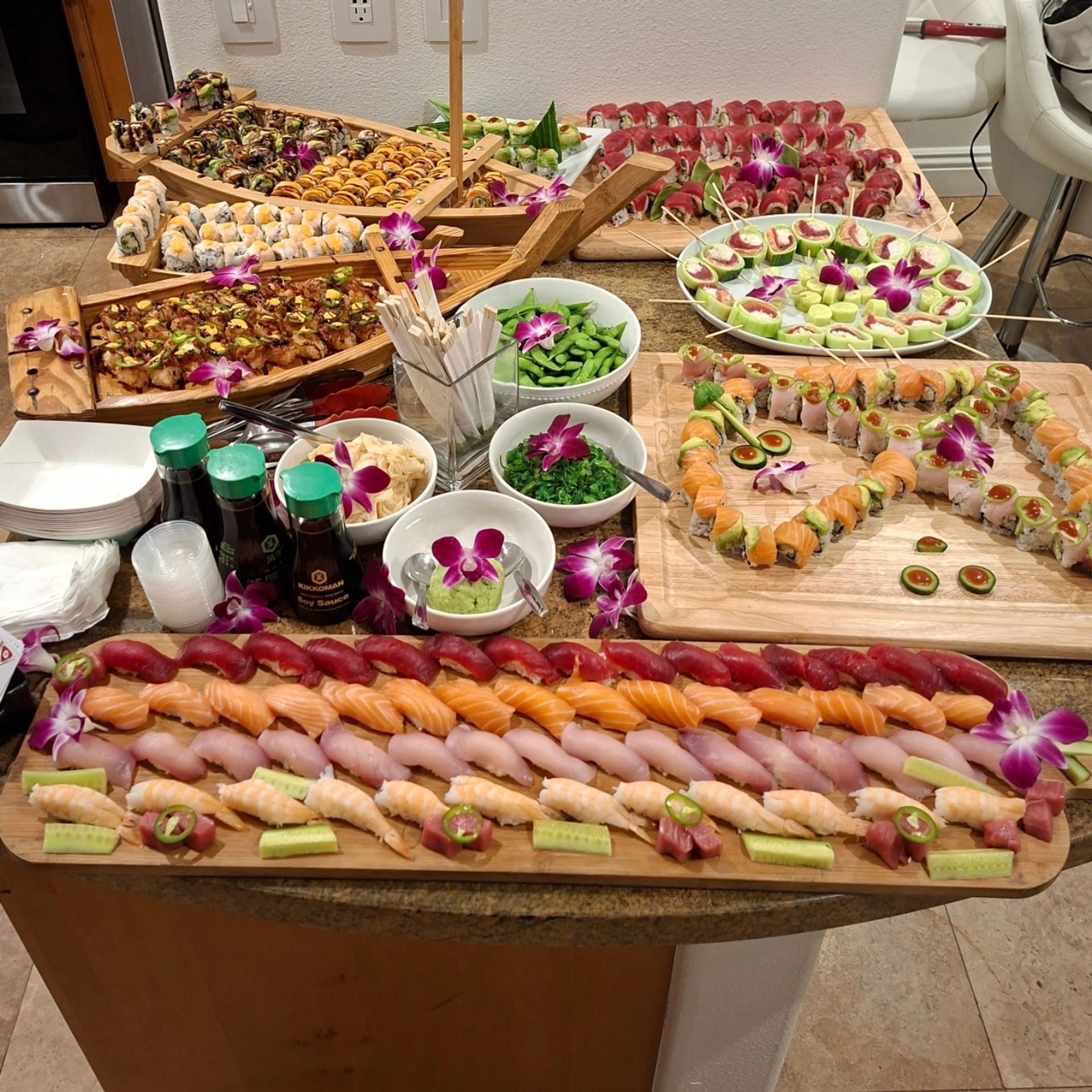 Vibrant sushi buffet and catering spread on wooden boards and boat platters with rows of nigiri, assorted maki rolls, cucumber bites, edamame, seaweed salad, wasabi and soy sauce bottles, orchid garnishes for a colorful party
