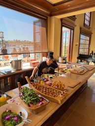 Chef preparing sushi, tempura and edamame buffet on a marble island in a sunlit waterfront kitchen with large windows overlooking a marina and outdoor diners