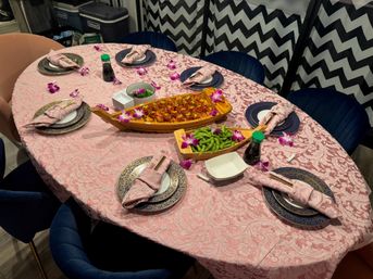 Oval dining table set for six with pink damask tablecloth, navy gold-rimmed plates, pink napkins with chopsticks, wooden sushi boats of seafood and edamame garnished with purple orchids and soy sauce bottles, chevron curtains in the background.