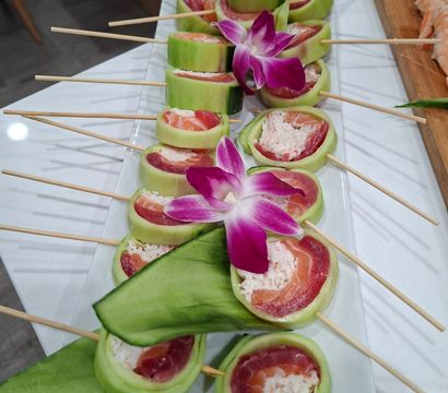 Cucumber-wrapped sushi skewers with tuna, salmon and crab filling, topped with purple orchid garnish on a white catering platter
