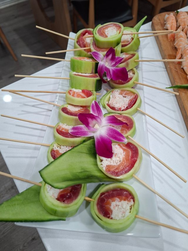 Cucumber-wrapped sushi rolls with salmon, tuna and crab filling on a white rectangular platter, bamboo skewers and purple orchid garnish
