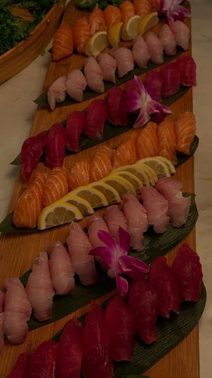 Assorted nigiri sushi — rows of salmon, tuna and white fish — neatly arranged on a long bamboo serving board with lemon slices, green leaves and purple orchid garnish.