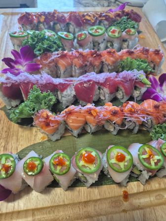 Assorted colorful sushi rolls on a wooden board — jalapeño-topped white fish, salmon and tuna-topped rolls with spicy sauces, purple orchid and kale garnishes.