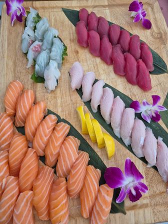 Vibrant sushi nigiri platter on a wooden board—rows of salmon, tuna and white-fish nigiri with lemon slices and purple orchid garnishes