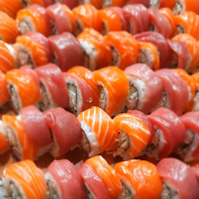 Close-up of fresh sushi rolls topped with alternating bright orange salmon and pink tuna slices, neatly arranged in rows on a platter