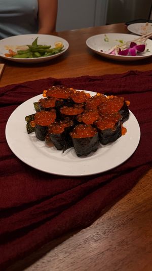 White plate of ikura (salmon roe) gunkan sushi—seaweed-wrapped rice topped with glossy orange roe—set on a burgundy cloth on a wooden dining table, with used plates, chopsticks and a drink in the background.