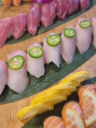 Sushi nigiri platter with rows of pink tuna and pale yellowtail topped with jalapeño slices, served with lemon and mandarin segments on green leaves over a wooden board