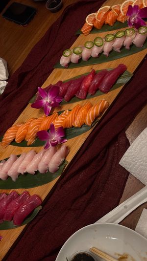 Assorted nigiri sushi platter on a long wooden board with rows of salmon, tuna and white fish topped with jalapeño and lemon slices, garnished with purple orchids, soy sauce and chopsticks on a table.