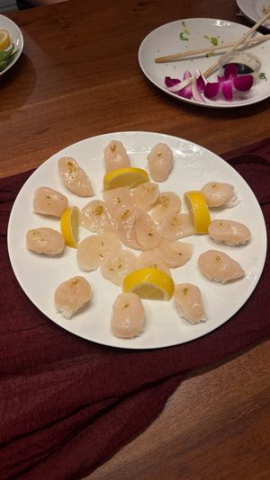 White plate of scallop nigiri sushi arranged in a ring with lemon wedges on a wooden dining table, chopsticks, soy sauce and edamame in the background