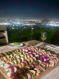 Vibrant sushi roll platter on a wooden board, garnished with purple orchids, with a wine glass and chopsticks on an outdoor table overlooking a twinkling city skyline at night.