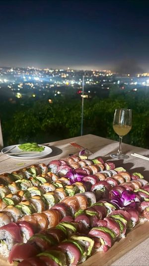 Vibrant sushi roll platter on a wooden board, garnished with purple orchids, with a wine glass and chopsticks on an outdoor table overlooking a twinkling city skyline at night.
