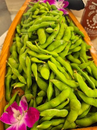 Close-up of bright green edamame pods piled in a wooden tray, lightly salted and garnished with a purple orchid.