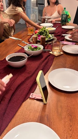 People gathered at a wooden dining table with a burgundy runner, empty plates, bowls of edamame, seaweed salad and soy sauce, drinks and a smartphone propped up for a casual dinner party.