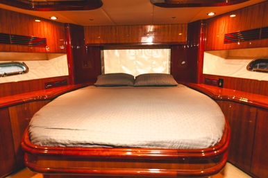 Cozy luxury yacht master cabin with glossy wood-paneled interior, rounded queen bed with pillows, warm recessed lighting and small porthole windows.