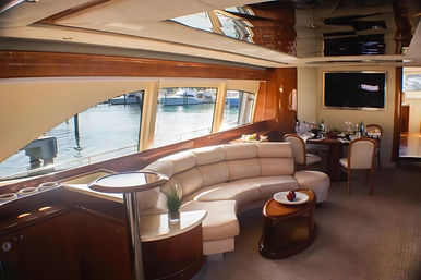Luxury yacht salon with curved cream leather sofa, glossy wood cabinetry, small dining table, flat-screen TV and large windows framing a marina water view