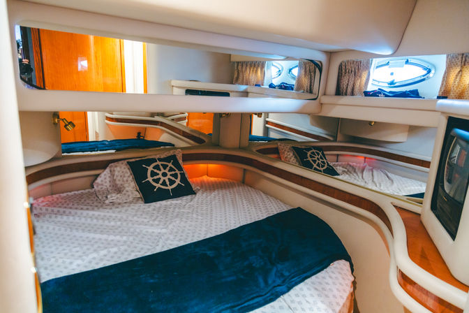 Cozy yacht cabin with rounded V-berth bed, nautical wheel pillow, navy throw, mirrored walls, wood trim, porthole window and warm ambient lighting