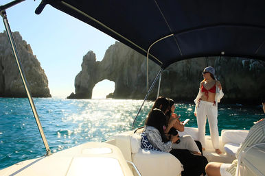 Friends on a motorboat in turquoise water approaching the sunlit rock arch at Cabo San Lucas, woman in a red bikini top and white pants standing under the boat canopy holding a drink