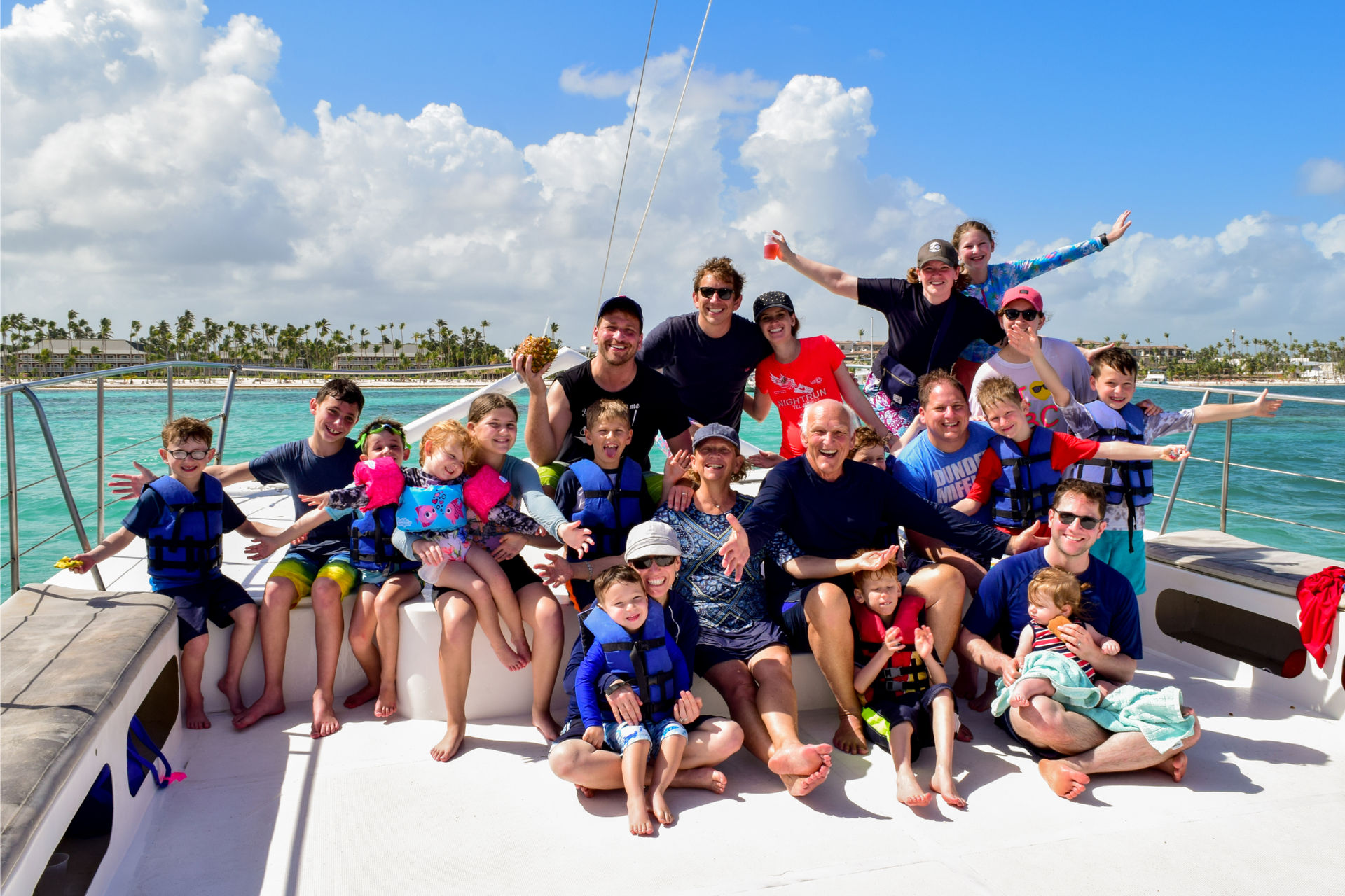 Multigenerational family on a catamaran cruise — smiling adults and children in life jackets on deck with turquoise ocean and palm-lined tropical shoreline under a sunny blue sky.