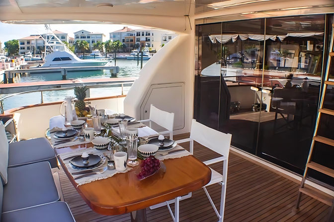Luxury yacht dining area on teak deck set for breakfast with tableware, fruit and chairs, overlooking a sunny marina with boats and waterfront condos