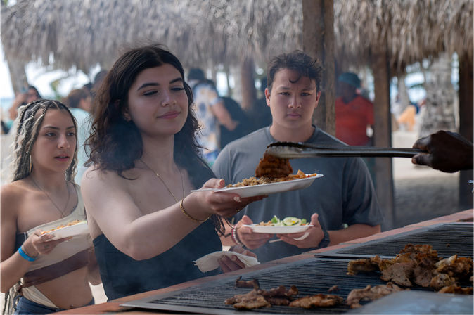 Beachside barbecue at a tropical palapa: young adults in line holding plates as a server uses tongs to add grilled meat from a large outdoor grill.