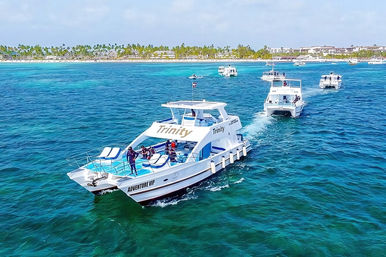 White tour catamaran with passengers cruising clear turquoise tropical waters near a palm-lined sandy beach, with several other excursion boats nearby under a bright blue sky