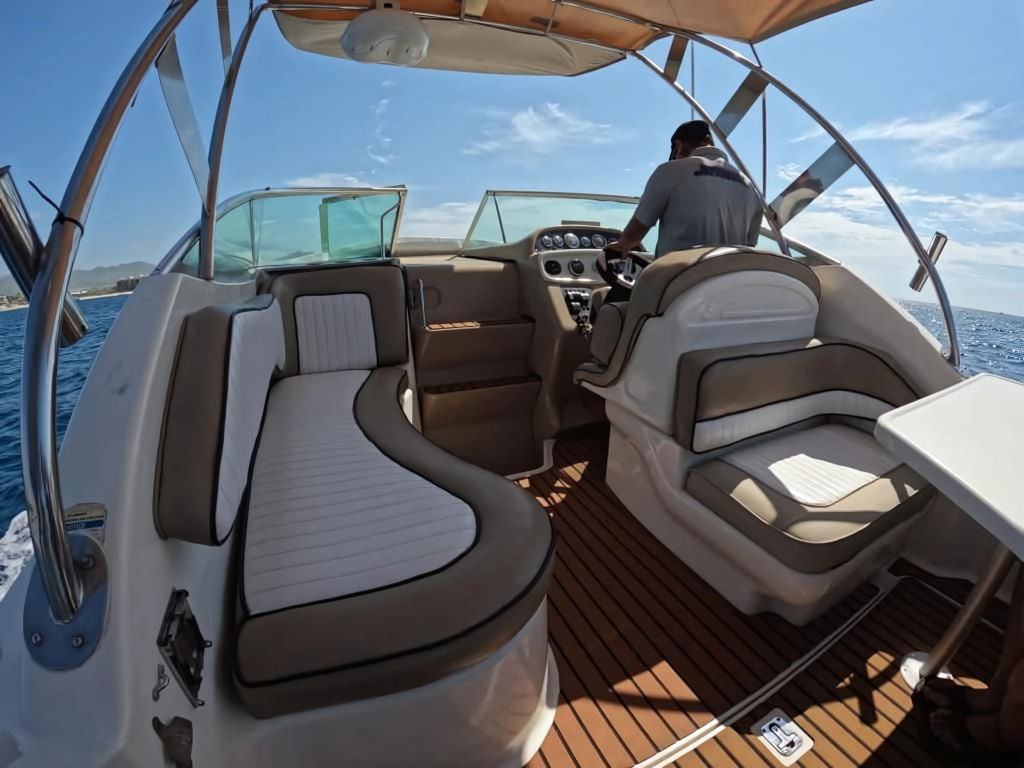 Spacious white-and-tan motorboat cockpit with cushioned seating and a person at the helm, cruising on bright blue coastal waters under a sunny sky.
