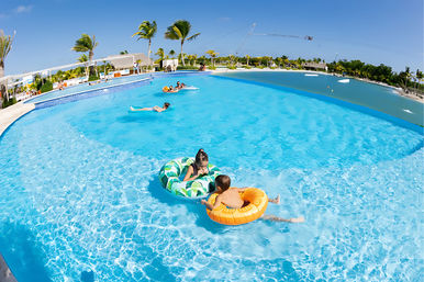 Kids floating on colorful inflatable tubes in a bright blue tropical resort pool, surrounded by palm trees, lounge chairs and a clear sunny sky — relaxed vacation scene.