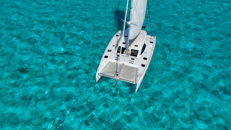 Aerial view of a white catamaran gliding over crystal-clear turquoise tropical water with sunlit ripples revealing the shallow seabed.