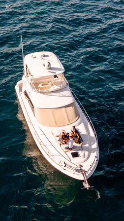 Aerial shot of a white motor yacht on deep blue ocean waters with a group of people lounging on the sunlit foredeck, relaxed boating scene.