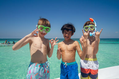 Three boys in colorful swim trunks and snorkeling masks pose on a sunny tropical beach with clear turquoise water, smiling and flashing playful hand signs.