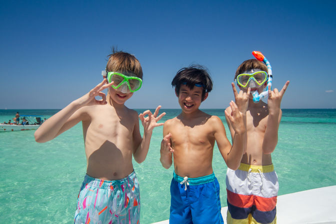 Three boys in colorful swim trunks and snorkeling masks pose on a sunny tropical beach with clear turquoise water, smiling and flashing playful hand signs.