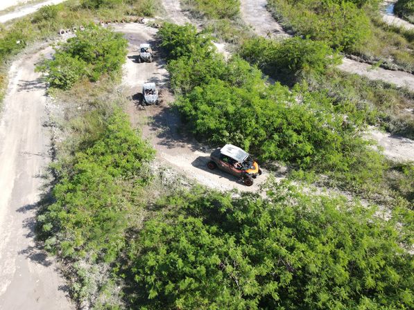 Aerial drone view of three off-road UTVs (side-by-sides) driving along a winding dirt trail through dense green scrubland and gravel paths.