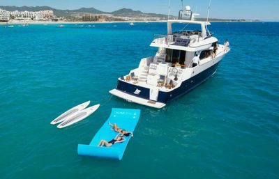 Luxury motor yacht anchored in turquoise sea off a resort-lined coastline, two people lounging on a bright blue floating mat with two paddleboards tied nearby on a sunny day