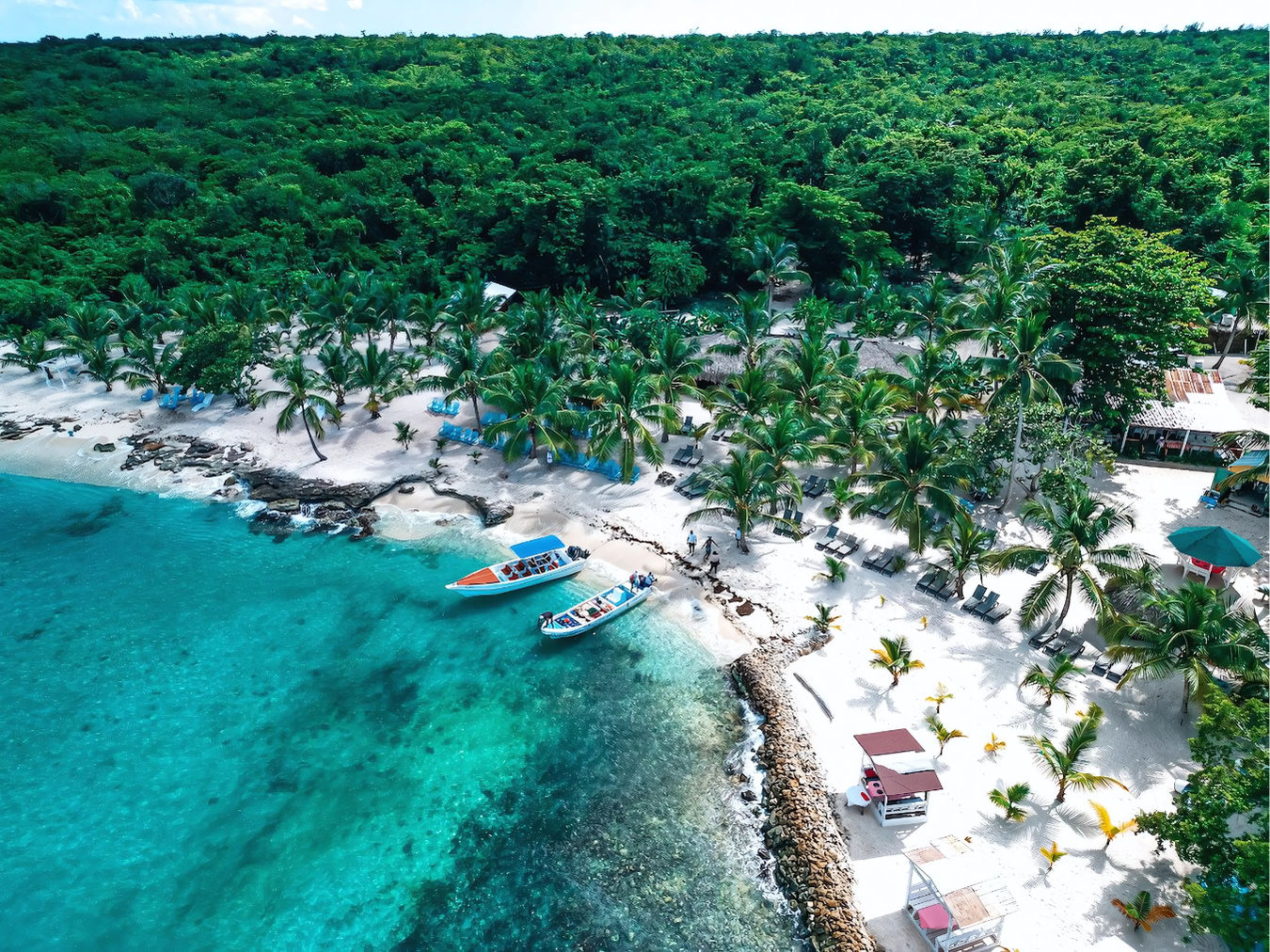 Aerial view of a tropical white-sand beach with turquoise water, palm trees and lounge chairs along the shore, two colorful motorboats anchored by rocky outcrops and dense green jungle behind the coast.