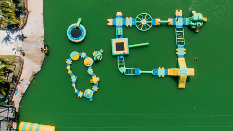 Aerial view of a colorful inflatable water park obstacle course on green lagoon water beside a palm-lined tropical shoreline, with slides, trampolines and a jet ski.